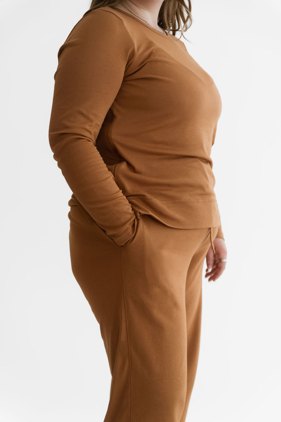 A person wearing a matching rust-brown knit lounge set with a long-sleeve top and pants, shown in side profile against a clean white studio background. - Amber